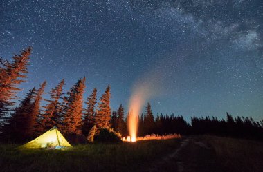 Yıldızlarla dolu gökyüzünün altında ve dağlarda Samanyolu 'nda gece kampı. Ormanın yakınındaki tepelerdeki aydınlık turist çadırlarının üzerinde yıldızlı gökyüzü. Gecenin karanlığında kamp ateşinden gelen sıcak ışık.