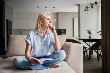 Genç bayan düşünceli bir şekilde başka tarafa bakıyor. Güzel bir kadın kanepede dinleniyor ve kitap okuyor. Günlük kıyafetlerle kanepede oturan ve düşünen bir kadın..