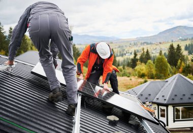 Evin çatısına fotovoltaik güneş modülü istasyonu kuranlar. Miğferli elektrikçiler güneş paneli sistemi kuruyor. Alternatif ve yenilenebilir enerji kavramı.