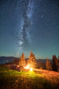 Yıldızlı gökyüzünün altındaki dağlarda gece kampı. Samanyolu ile dolu gökyüzünün altındaki kamp alanında turist çadırları. Yürüyüşçülerin silueti yanan kamp ateşinin yanında dinleniyor. Turizm kavramı, maceralar.