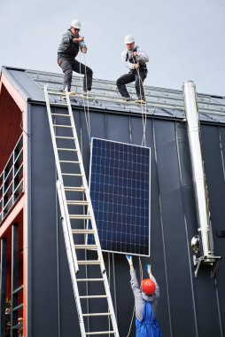 Erkek çatıcılar evin çatısına güneş paneli kuruyor. Miğferli teknisyenler fotovoltaik güneş modülünü açık havada halatlarla kaldırıyorlar. Alternatif ve yenilenebilir enerji kavramı.