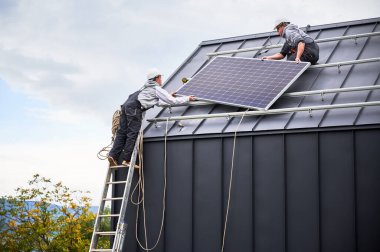 Atçılar evin çatısına güneş paneli inşa ediyorlar. Dışarıda fotovoltaik güneş modülü taşıyan miğferli işçiler. Alternatif ve yenilenebilir enerji kavramı.