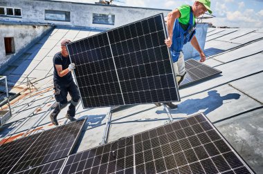 İşçiler evin metal çatısında güneş paneli inşa ediyorlar. Dışarıda fotovoltaik güneş modülü taşıyan iki adam. Alternatif, yeşil ve yenilenebilir enerji üretimi kavramı.