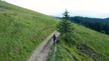 Dışarıda elektrikli dağ bisikleti süren bisikletçi. Erkek turistin dağlardaki otlak patika boyunca bisiklet sürüşünü, kaskını ve sırt çantasını gösteren hava manzarası. Spor anlayışı, aktif eğlence ve doğa.