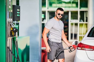 Benzin deposuna benzin dolduran bir adamın portresi. Kendine güvenen sürücü uzun bir yol için tam depo dolduruyor. Benzin istasyonunun arka planında yetişkin bir erkek yakıt deposu var..