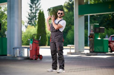 Güçlü işçi yakıt dolu bir teneke kutuyu omuzunda tutuyor. Kendine güvenen bir benzin istasyonu operatörü, gaz dolu ağır sarnıçları umursuyor. Güneş gözlüklü ve tulumlu erkek işçi benzin istasyonunun arka planında duruyor..