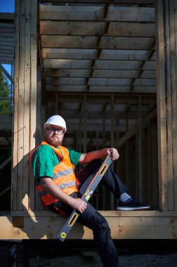 Marangoz ahşap çerçeve iki katlı ev inşa ediyor. Sakallı, gözlüklü, gözlüklü koruyucu kask takan, tulumlu ve turuncu yelek giyen bir adam. Modern ekolojik yapı kavramı.