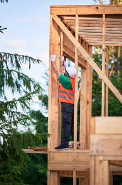 Marangoz ormanın yakınında iki katlı ahşap çerçeve inşa ediyor. Adam koruyucu kask ve inşaat yeleği giyerken çivi çakıyor. Modern ekolojik yapı kavramı