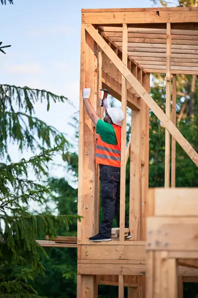 Marangoz ormanın yakınında iki katlı ahşap çerçeve inşa ediyor. Adam koruyucu kask ve inşaat yeleği giyerken çivi çakıyor. Modern ekolojik yapı kavramı
