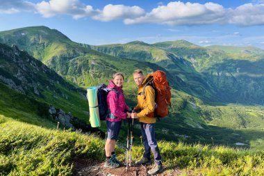 İki yürüyüşçünün portresi, turuncu sırt çantalı bir adam ve yeşil yastıklı pembe ceketli bir kadın, dağ yolunda duruyorlar. Arka planda parlak mavi gökyüzünün altındaki göz kamaştırıcı yeşil tepeler.