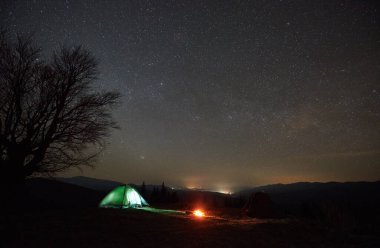 Night camping. Lone tent glows softly beside campfire under star-studded sky. Silhouette of leafless tree stands against mountainous horizon, creating serene and mesmerizing night scene in nature. clipart