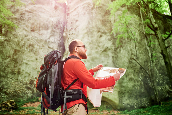 Bearded tourist man studies map while stands in front of large rock in dense forest. Traveler with grey backpack and glasses, planning outdoor adventure, hiking or climbing route.