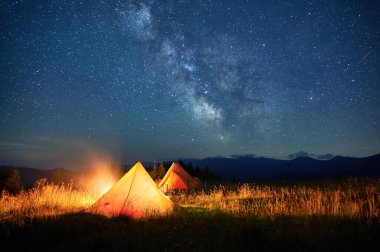 Yıldızlarla dolu parlak gökyüzünün altında, çimenli yamaçlarda parlayan iki çadır. Samanyolu belirgin bir şekilde görülebilir. Sıcak kamp ateşi doğadaki berrak gecenin dingin güzelliğini aydınlatan manzarayı aydınlatır..