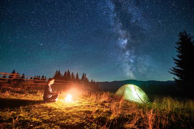 Kamp ateşinin yanında dinlenen yürüyüşçü, elinde fincanla baş döndürücü yıldızlı gökyüzünün altında parlayan bir çadır tutuyor. Samanyolu yukarıda ışıl ışıl parlıyor. Huzurlu dağ ortamında büyülü ve huzurlu bir ortam yaratıyor..