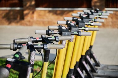Sarı gidonu düzgün bir şekilde kaldırıma dizilmiş elektrikli scooterlar. Yakın plan görünümü, kolay kiralama ve kullanım için çeşitli kontrol düğmelerini ve QR kodlarını gösteren gidonlara odaklanır.