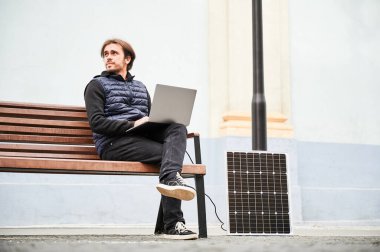 Adam ahşap bankta oturuyor ve güneş paneline bağlı dizüstü bilgisayarla çalışıyor. Kentsel ortam ve günlük giysiler modern, sürdürülebilir, mobil, çevre dostu çalışma alanını vurguluyor.