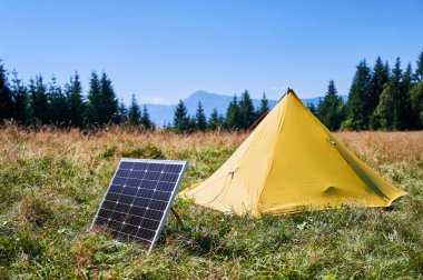 Çimenli alana kurulmuş güneş paneli olan sarı çadır açık mavi gökyüzünün altında sık ve yeşil ağaçlarla çevrili. Çevre dostu kamp, yenilenebilir enerjinin açık hava aktivitelerine entegrasyonu.