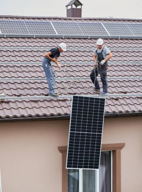 Erkek yükleyiciler fotovoltaik güneş modüllerini evin çatısına kaldırıyorlar. Kasklı elektrikçiler dışarıda güneş paneli sistemi inşa ediyorlar. Alternatif ve yenilenebilir enerji kavramı.