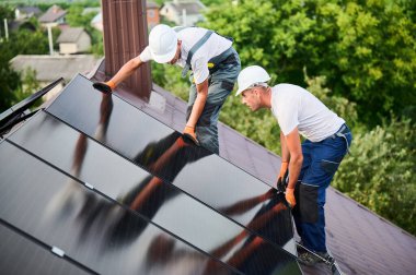 İşçiler evin çatısına güneş paneli inşa ediyorlar. Dışarıda fotovoltaik güneş modülü kuran kask takan iki adam. Alternatif, yeşil ve yenilenebilir enerji üretimi kavramı.