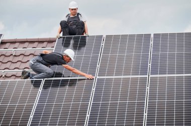 Erkek montajcılar fotovoltaik güneş modüllerini evin çatısına monte ediyorlar. Kasklı elektrikçiler dışarıda güneş paneli sistemi kuruyorlar. Alternatif ve yenilenebilir enerji kavramı.