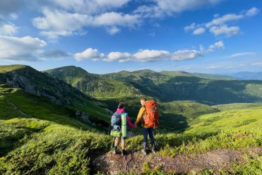 El ele tutuşan iki yürüyüşçünün arka görüntüsü dağ manzarasına bakan patikada duruyor. Sırt çantaları ve yürüyüş direkleriyle donatılmış turistler. Yuvarlanan tepeler, kayalık çıkıntılar, bulutlu parlak mavi gökyüzü.
