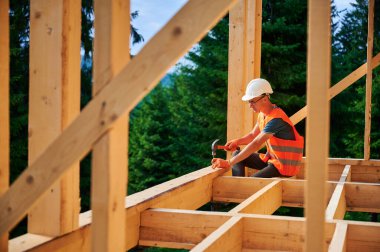 Marangoz tarafından inşa edilen ahşap çerçeve. Çekiç kullanarak çivi çakan gözlüklü bir adam koruyucu kask ve çelik yelek giyiyor. Modern ekolojik yapı kavramı.