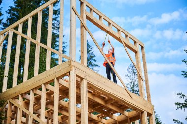 Marangoz iki katlı ahşap çerçeve dikiyor. Adam iş kıyafetleri ve kaskla mezura kullanarak ölçü alıyor. Modern ekolojik yapı kavramı.