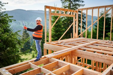 İşçi, ormanın yakınında iki katlı ahşap bir bina inşa ediyor. Tulum giymiş sakallı bir adam ve inşaat planlarını gözden geçiriyor. Modern, çevre dostu inşaat uygulamaları kavramı.