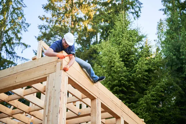 Ahşap işçisi, ormanın yakınında iki katlı çerçeve ev inşa ediyor. Sakallı, gözlüklü adam güvenlik kaskı takarken çivi çakıyor. Modern ekolojik yapı kavramı.