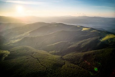 Gün batımında sakin dağ manzarasının havadan görüntüsü. Orman tepeleri sıcak, altın renginde ışıkla yıkanarak sakin bir atmosfer yaratıyor. Yoğun, yemyeşil yeşilliklerle kaplı yuvarlanan tepeler uzaklara kadar uzanıyor..
