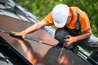 Evin çatısında fotovoltaik güneş paneli inşa eden bir işçi. Miğferlerde mühendis ve eldivenler dışarıda altıgen anahtarla güneş modülü kuruyor. Alternatif enerji üretimi kavramı.