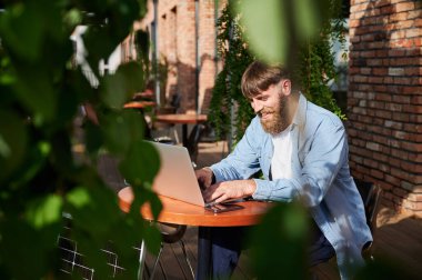 Adam dizüstü bilgisayarda, akıllı telefon da açık havada çalışıyor. Yakınlardaki güneş paneli, teknoloji ve sürdürülebilirliği birleştiren sürdürülebilir, modern çevre dostu çalışma alanını vurguluyor.