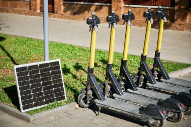 Kaldırımda çimlerin yanına park edilmiş sarı gidonu olan beş elektrikli scooter. Güneş paneli yakınlarda, scooterlar için çevre dostu sürdürülebilir şarj çözeltisi gösteriyor.