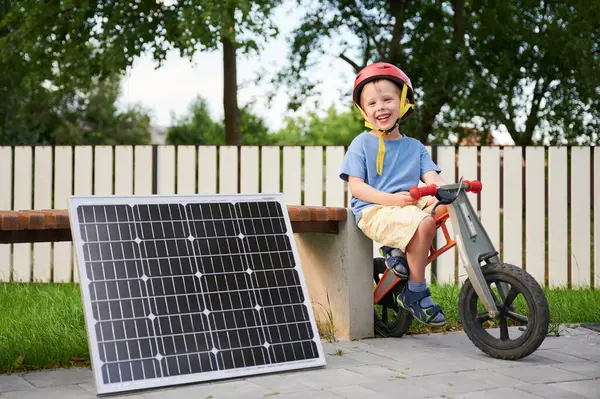 Stock image Young happy boy sits on balance bike next to solar panel and wooden bench, wearing red helmet, smiling brightly at camera. Background features white fence, trees. Concept of green energy generation.