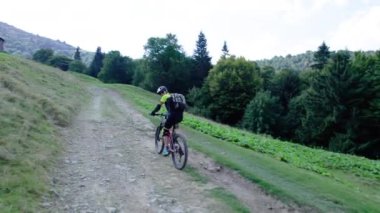 Dışarıda elektrikli dağ bisikleti süren bisikletçi. Erkek turist dağlarda otlak patika boyunca bisiklet sürüyor, başlık ve sırt çantası takıyor. Spor anlayışı, aktif eğlence ve doğa.