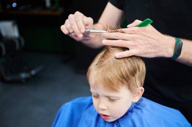 Erkek berber, tarak ve makas kullanarak küçük çocukların saçını kesiyor. Çocuğun saçlarını yetişkin bir erkekten kestiriyor, muhtemelen berber. Modern berber dükkanında profesyonel kuaför ve şirin bir müşteri..