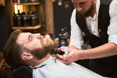 Profesyonel berber sakal tıraşı için elektrikli tıraş makinesi kullanıyor. Berber dükkanında düzenli bir müşteri. Kendine güvenen erkek müşteri sakalını yaptırıyor..