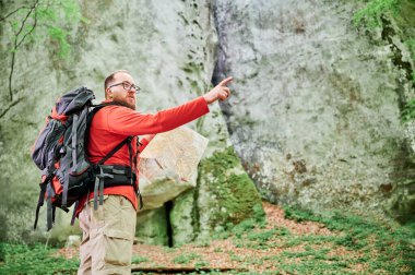 Sakallı turist, sık ormandaki büyük kayaların önünde dururken haritayı inceliyor. Gri sırt çantası ve gözlüklü gezgin, önünü gösteriyor, açık hava macerası planlıyor, yürüyüş ya da tırmanma rotası çiziyor..