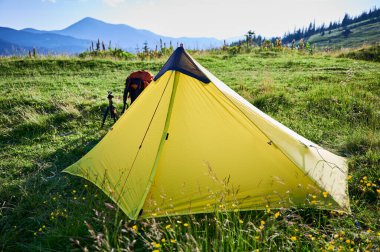 Çimenli yamaçta sarı turist çadırı. Kamp malzemeleri de yakınlarda. Sersemletici yeşil tepeler, berrak mavi gökyüzünün altındaki uzak dağlar, barışçıl açık hava maceraları için mükemmel bir yer yaratıyor..