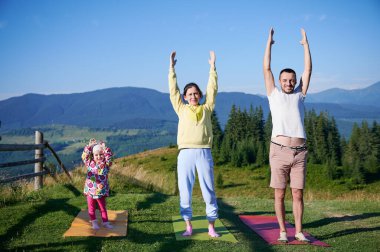 Genç bir aile sabahları doğa manzaralı dağlarda yoga yapıyor. Baba, anne ve kız yoga minderlerinde, arka planda dağları ve ormanı manzaralı açık mavi gökyüzünün altında.