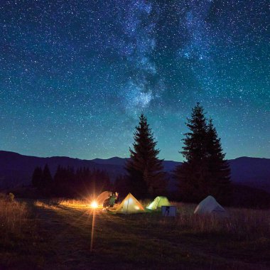 İki turist, kamp ateşi altında koltuklarda dinleniyor. Yanan çadırları, nefes kesen yıldızlı gökyüzünün altında. Samanyolu, dağ yabanda büyülü ve huzurlu bir atmosfer yaratarak yukarılara uzanır..