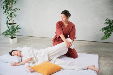 Masör Tayland yogası masajı yapıyor. Geleneksel masaj tedavisi uygulayan kadın terapist, kadın hastaların vücudunu geriyor. Müşteri paspasın üzerinde uzanıyor, sakin atmosferde el masajının keyfini çıkarıyor..