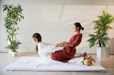 Masör Tayland yogası masajı yapıyor. Geleneksel masaj tedavisi uygulayan kadın terapist, kadın hastaların vücudunu geriyor. Müşteri paspasın üzerinde uzanıyor, sakin atmosferde el masajının keyfini çıkarıyor..
