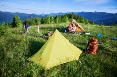 Çimenli tepenin üzerinde çadırları olan resimli bir dağ kampı. Kampçı ve çocuk kamp ateşine eğilimli, sırt çantaları ve teçhizatlarla çevrili. Yuvarlanan tepeler, yemyeşil, açık mavi gökyüzü. Barışçıl açık hava macerası.