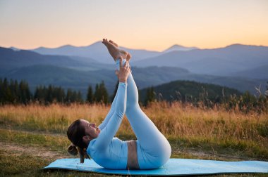 Dağlarda yoga yapan bir kadın, sakin, doğal bir ortamda. Mavi hasırda yoga yapan kadın, gün doğumunda ya da günbatımında güzel dağ manzarasının arkaplanıyla.