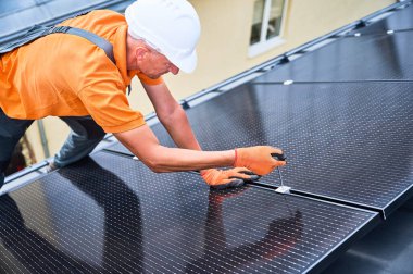 Evin çatısında fotovoltaik güneş paneli inşa eden bir işçi. Miğferlerde mühendis ve eldivenler dışarıda altıgen anahtarla güneş modülü kuruyor. Alternatif enerji üretimi kavramı.