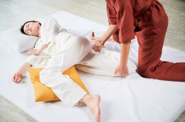 Masör Tayland yogası masajı yapıyor. Geleneksel masaj tedavisi yapan kadın terapistin kadın hasta vücudunu esnetmesi. Müşteri paspasın üzerinde uzanıyor, sakin atmosferde el masajının keyfini çıkarıyor..