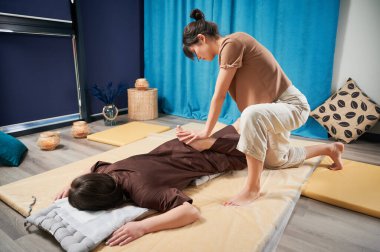 Masör Tayland yogası masajı yapıyor. Geleneksel masaj tedavisi uygulayan kadın terapist, kadın hastaların vücudunu geriyor. Müşteri paspasın üzerinde uzanıyor, sakin atmosferde el masajının keyfini çıkarıyor..