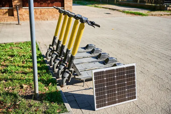 stock image Row of five electric scooters with yellow handlebars parked on sidewalk next to patch of grass. Solar panel placed nearby, indicating eco-friendly sustainable charging solution for scooters.