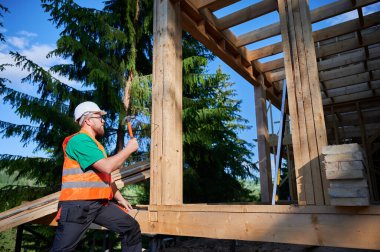 Marangoz ormanın yakınında ahşap çerçeve inşa ediyor. Sakallı adam koruyucu kask ve inşaat yeleği giyerken çivi çakıyor. Modern ekolojik yapı kavramı.